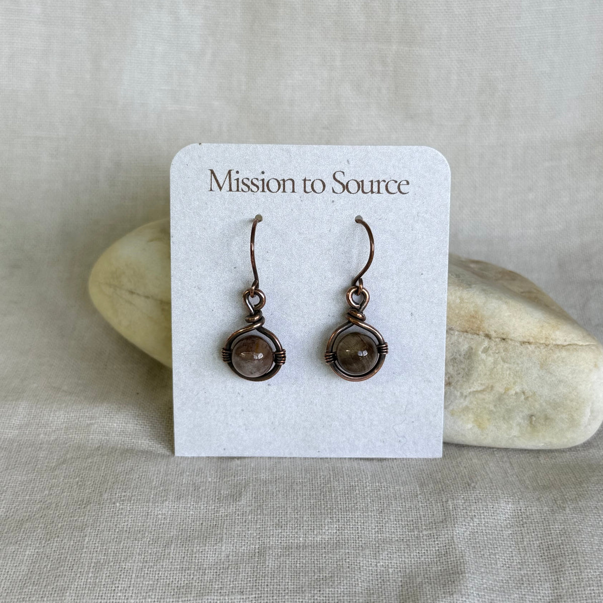 Pair of copper earrings with brown gemstones on a 'Mission to Source' card with a stone background