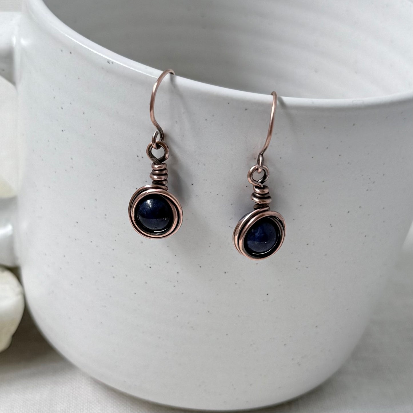 A pair of handmade copper wire-wrapped drop earrings with dark blue round gemstones, shown hanging from the rim of a white speckled ceramic mug.