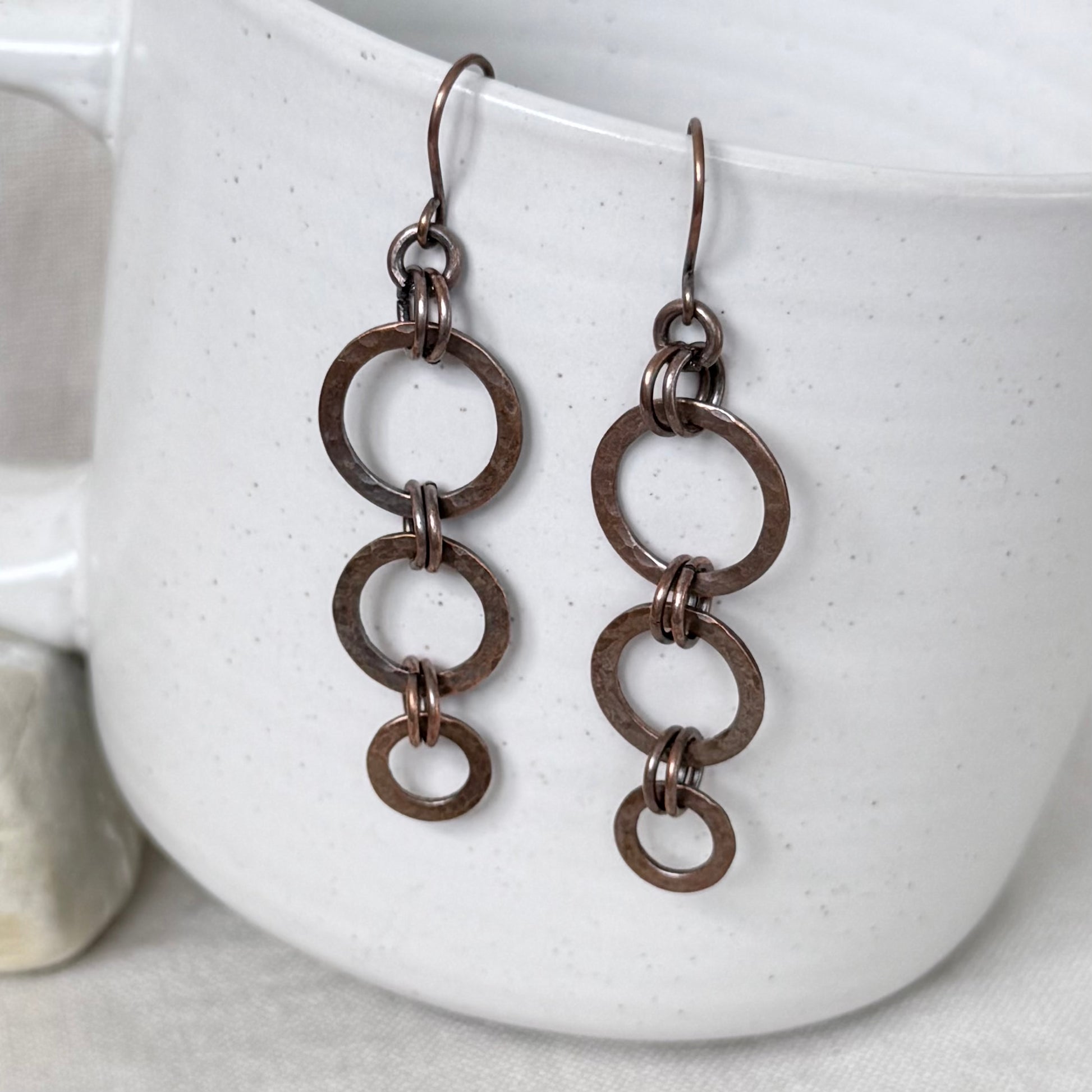 A pair of copper drop earrings with three stacked circular links, hanging from hooks against a white speckled ceramic mug.