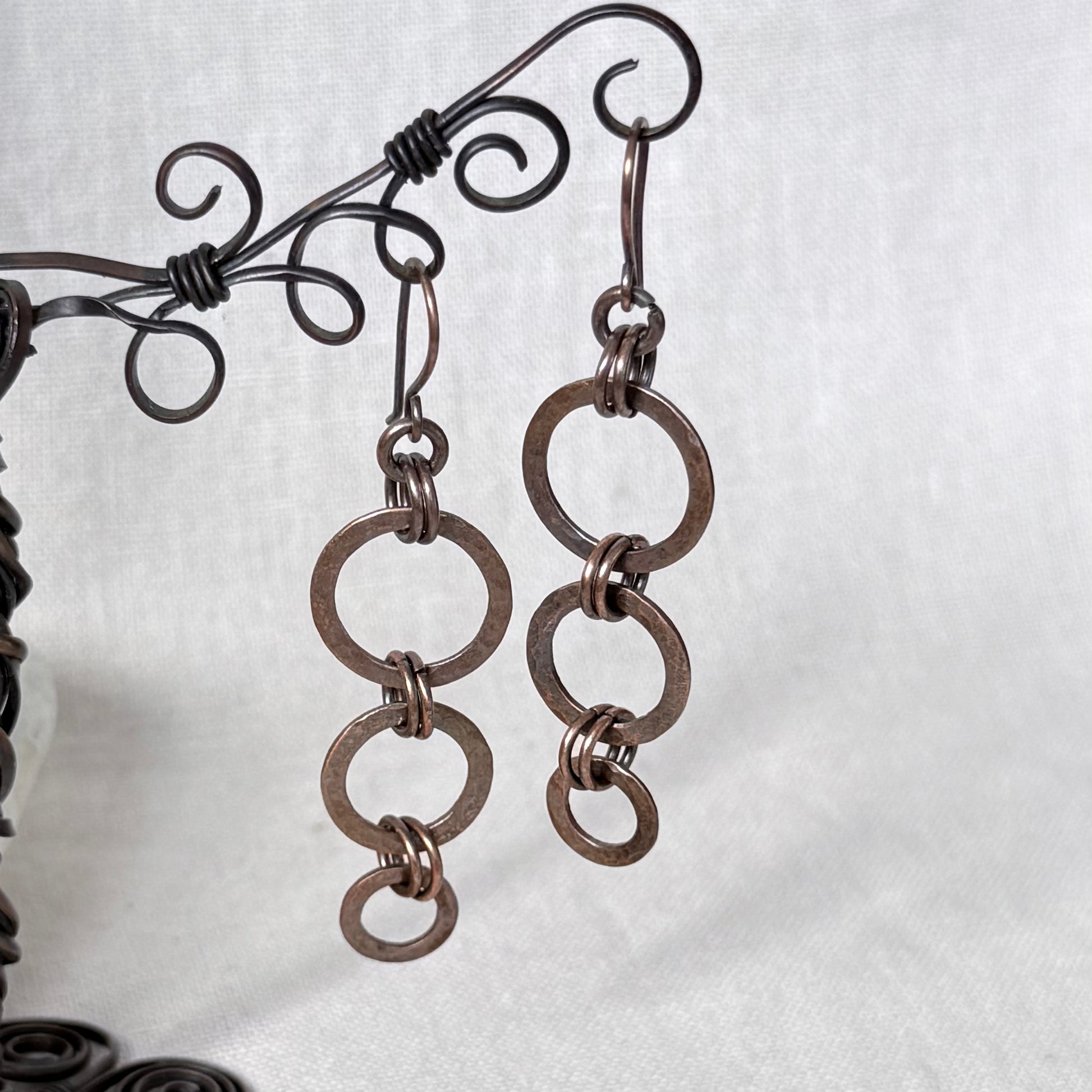 A pair of handmade copper dangle earrings made of interlinked circular wire links, hanging from a decorative dark metal wire display stand against a light grey fabric background.