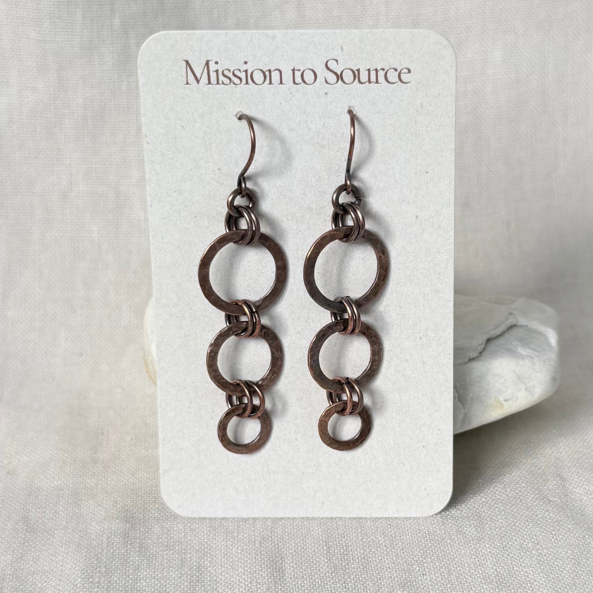 A pair of copper dangle earrings with interlocking circular links, displayed on a white card labeled 'Mission to Source' against a soft neutral fabric background.