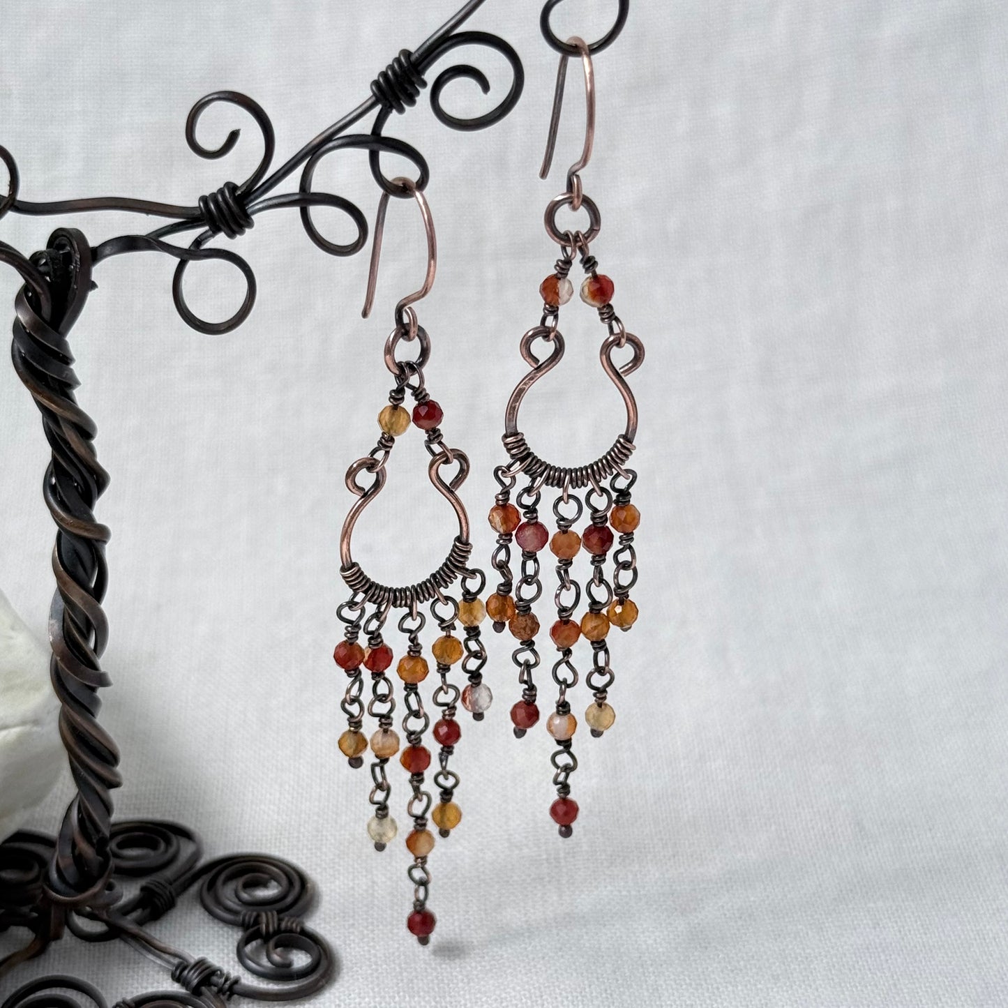 A pair of copper chandelier drop earrings with orange Carnelian beads, displayed on a black metal wire stand against a light grey fabric background.