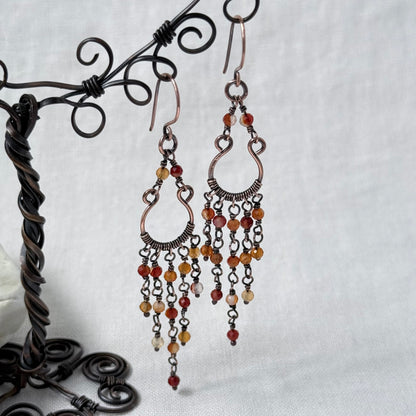 A pair of copper chandelier drop earrings with orange Carnelian beads, displayed on a black metal wire stand against a light grey fabric background.
