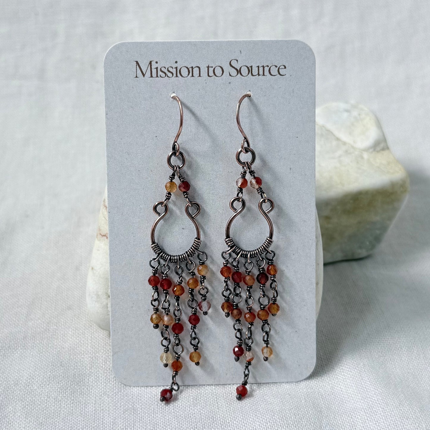 A pair of dangling copper earrings displayed on a white card labeled 'Mission to Source.' The earrings have multiple dangling chain strands set with small orange and red carnelian beads.