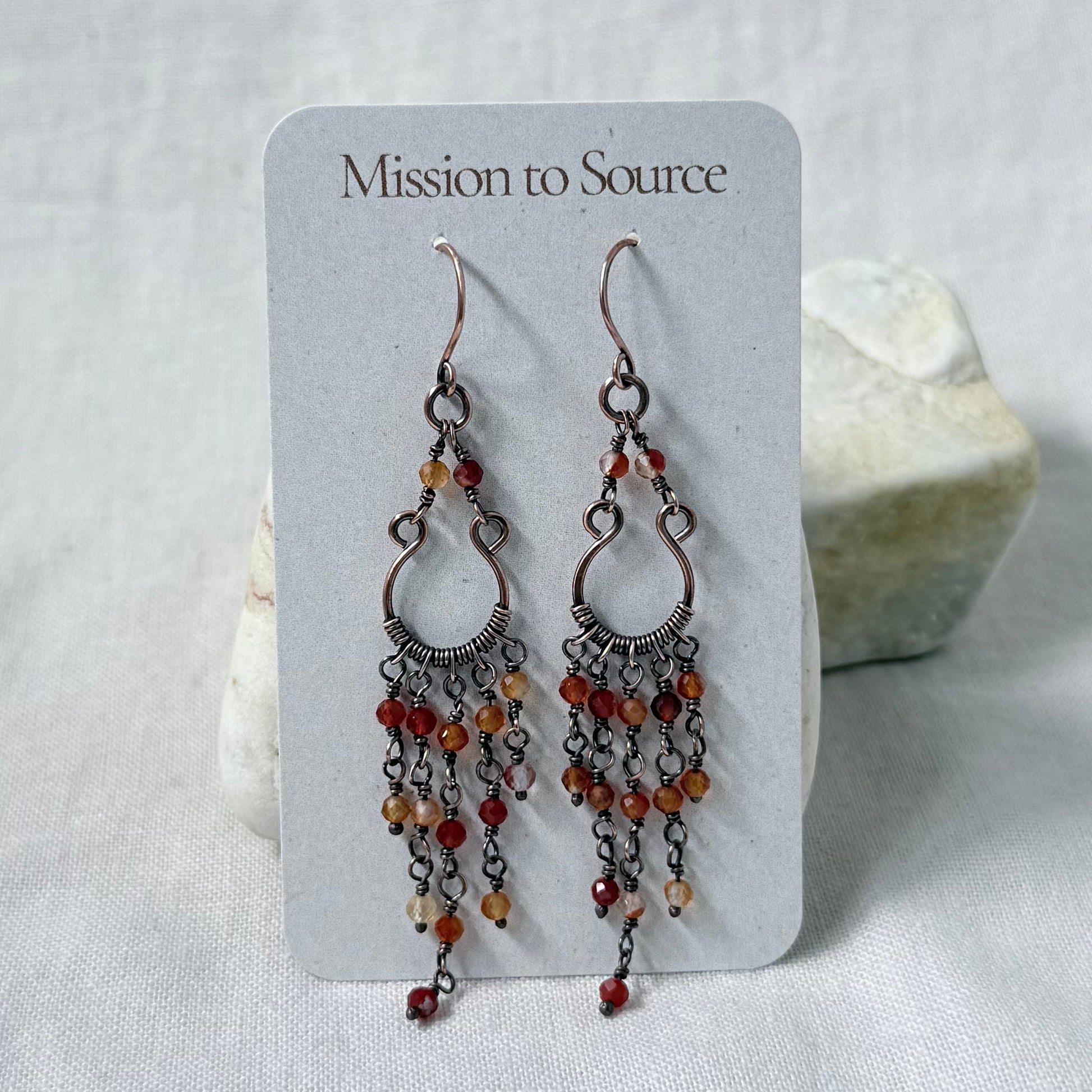 A pair of dangling copper earrings displayed on a white card labeled 'Mission to Source.' The earrings have multiple dangling chain strands set with small orange and red carnelian beads.