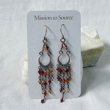 A pair of dangling copper earrings displayed on a white card labeled 'Mission to Source.' The earrings have multiple dangling chain strands set with small orange and red carnelian beads.