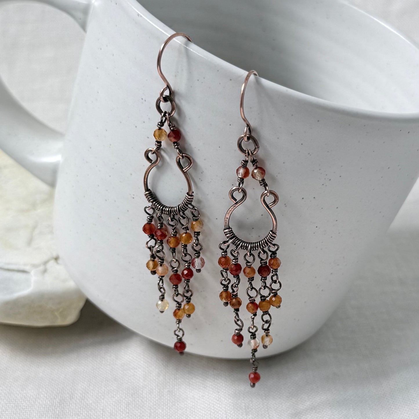 A pair of dangling copper chandelier earrings with orange toned Carnelian beads, displayed against a white ceramic mug.