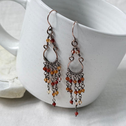 A pair of dangling copper chandelier earrings with orange toned Carnelian beads, displayed against a white ceramic mug.
