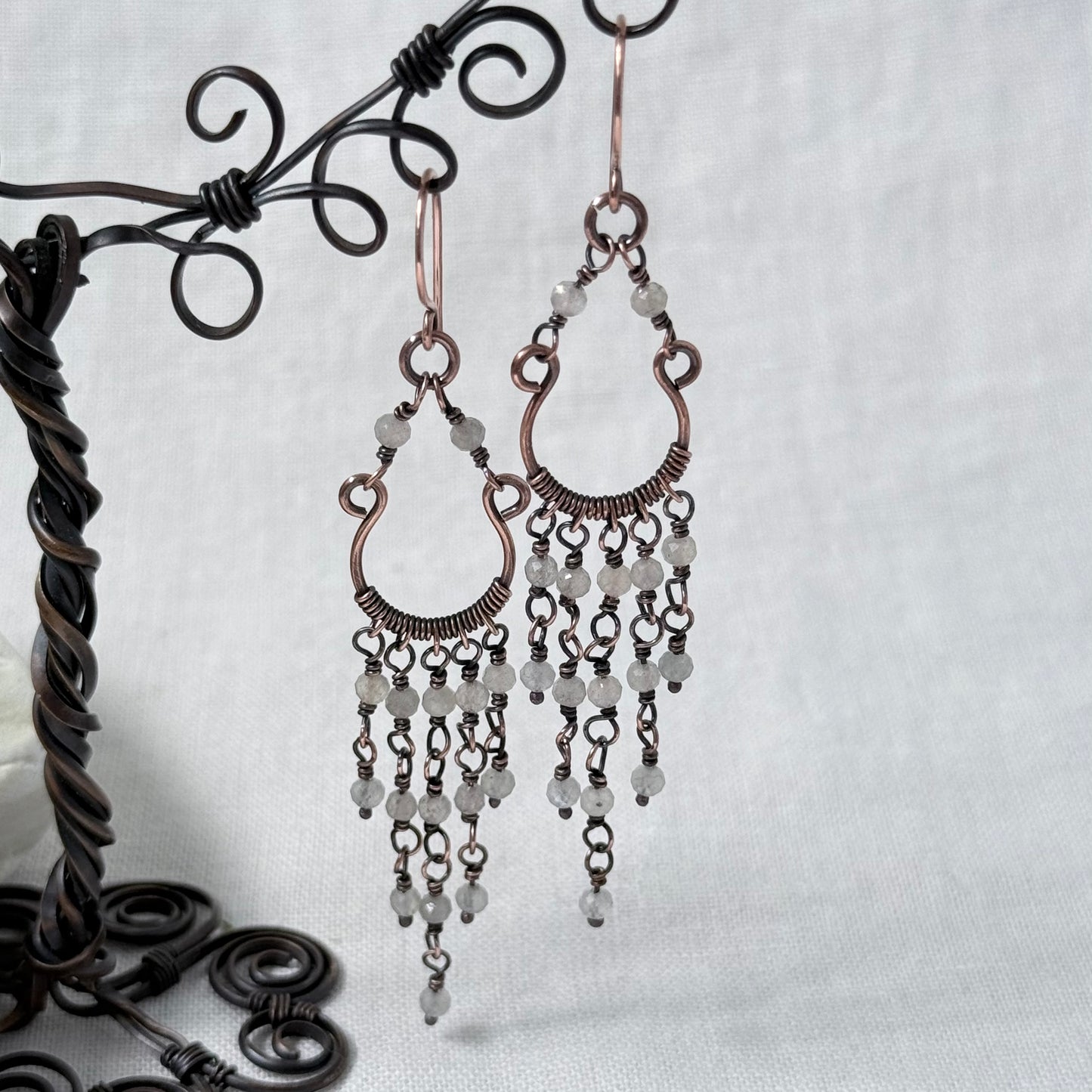 A pair of copper chandelier drop earrings with white Labradorite beads, displayed on a black metal wire stand against a light grey fabric background.