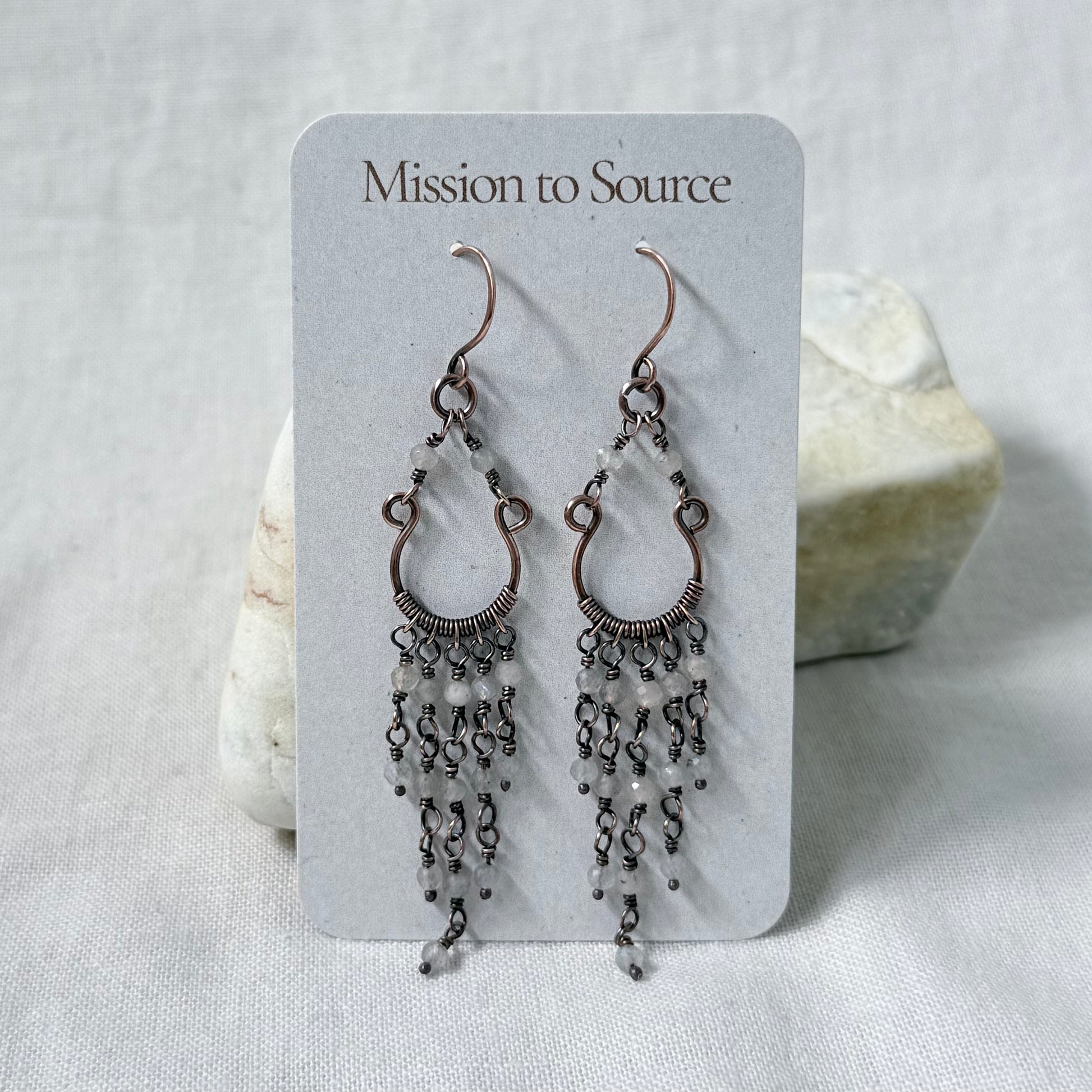 A pair of dangling copper chandelier earrings with pale Labradorite beads, displayed on a white card labeled 'Mission to Source,' set against a soft white fabric background with a small stone prop.