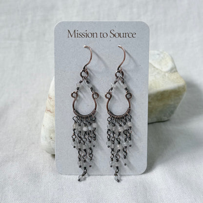 A pair of dangling copper chandelier earrings with pale Labradorite beads, displayed on a white card labeled 'Mission to Source,' set against a soft white fabric background with a small stone prop.