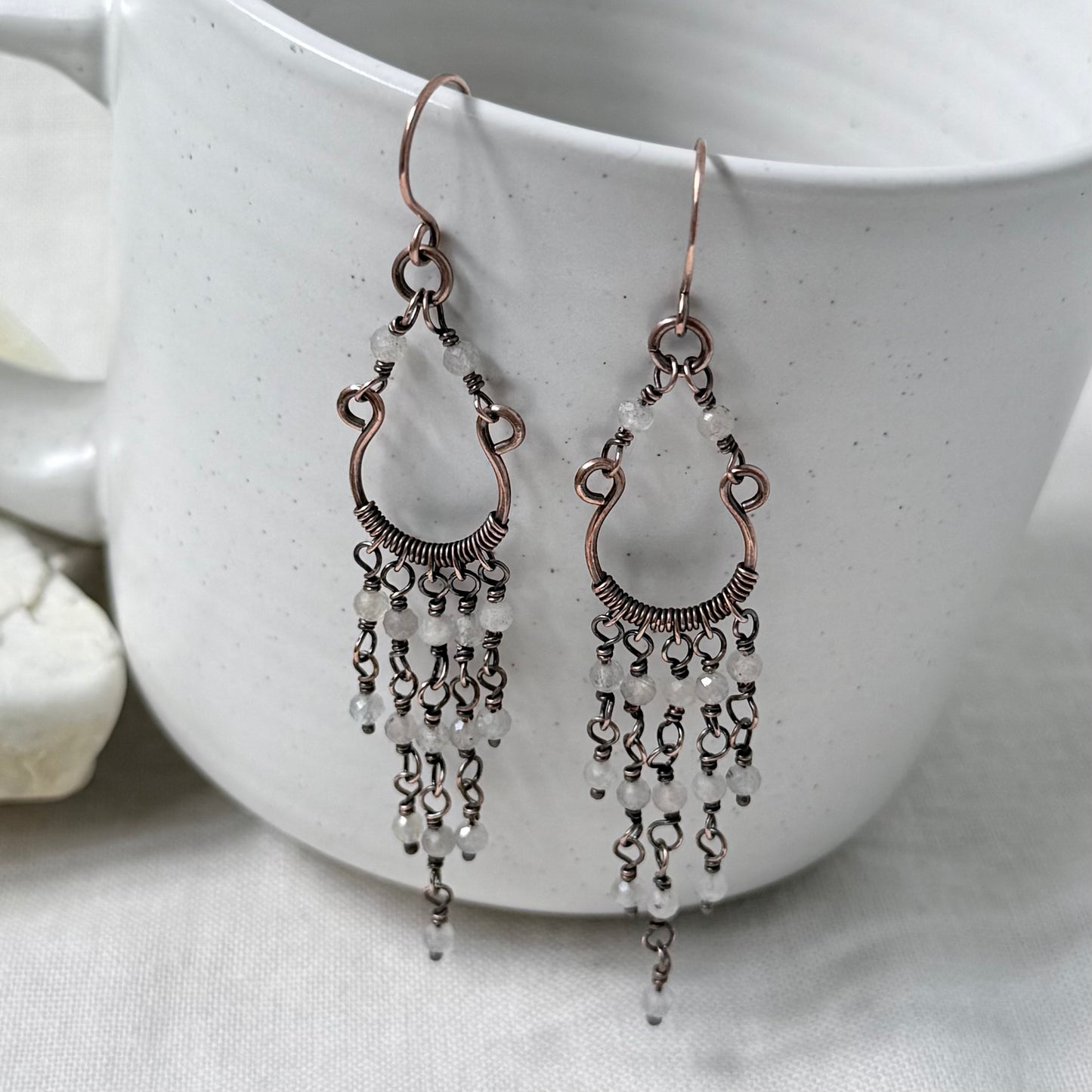 A pair of copper chandelier dangle earrings, textured wire detailing, and cascading pale Labradorite faceted beads, displayed on a white ceramic cup.