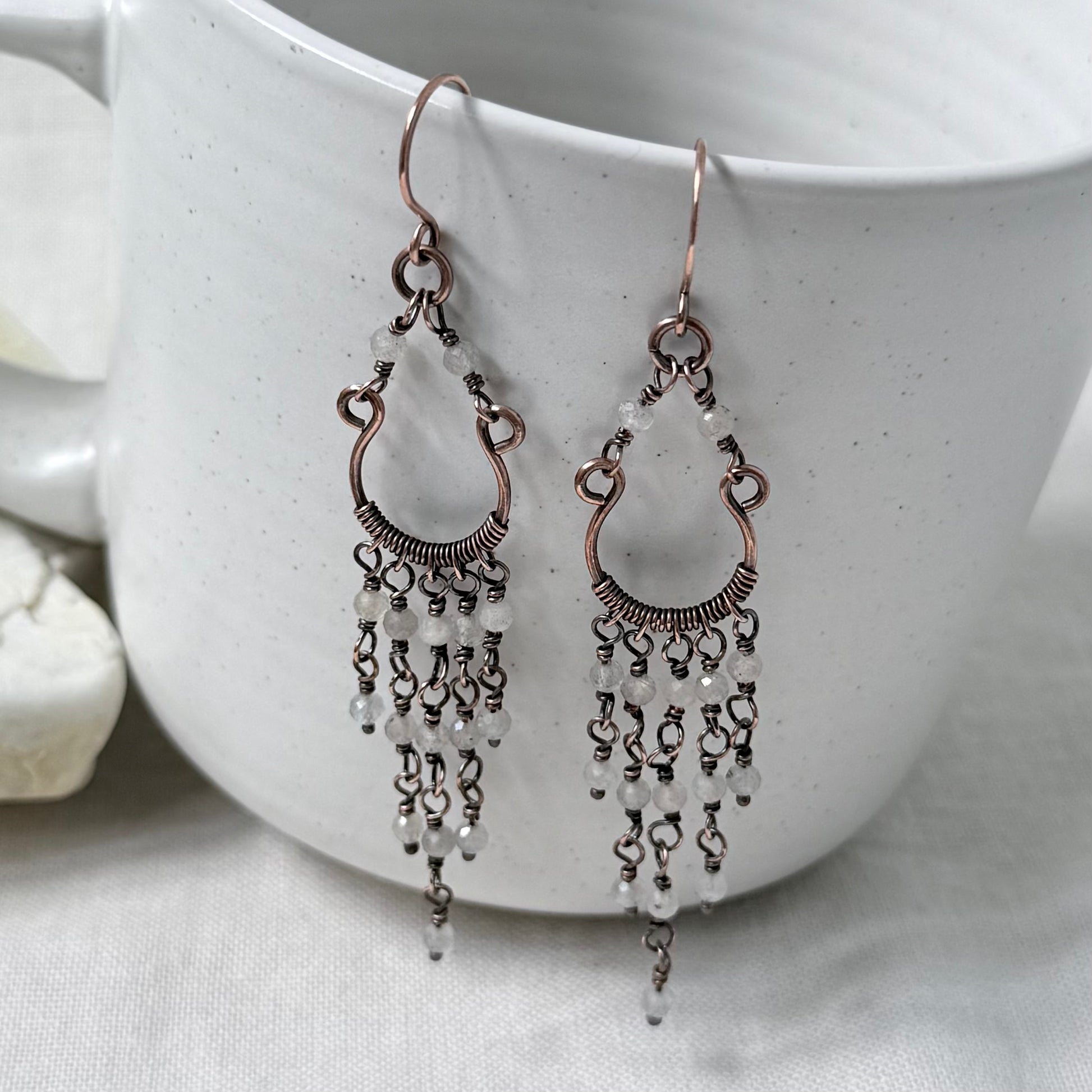A pair of copper chandelier dangle earrings, textured wire detailing, and cascading pale Labradorite faceted beads, displayed on a white ceramic cup.