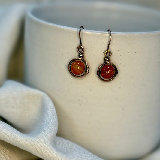 Pair of Copper Earrings with a round orange Carnelian Bead hanging from a cream coloured mug on linen background