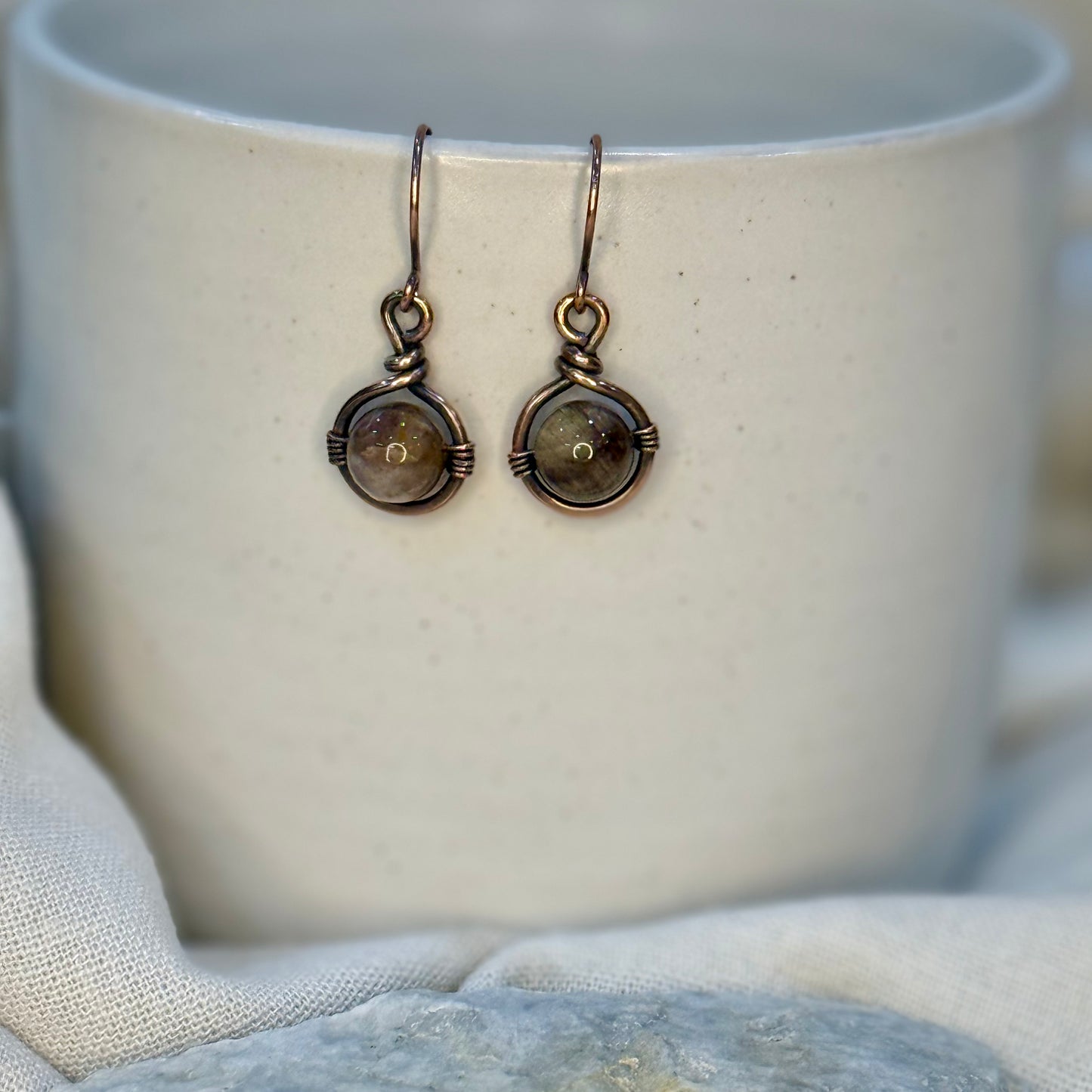 Pair of Copper Earrings with a round brown petrified wood Bead hanging from a cream coloured mug on linen background