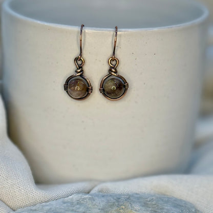 Pair of Copper Earrings with a round brown petrified wood Bead hanging from a cream coloured mug on linen background