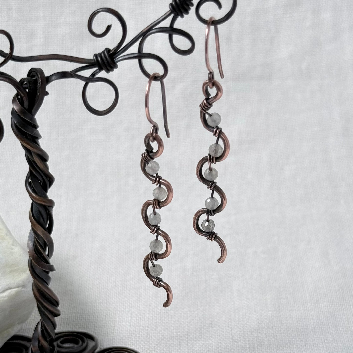 A pair of handmade copper wire drop earrings with small Labradorite faceted beads, displayed on a dark metal wire stand against a light grey fabric background.