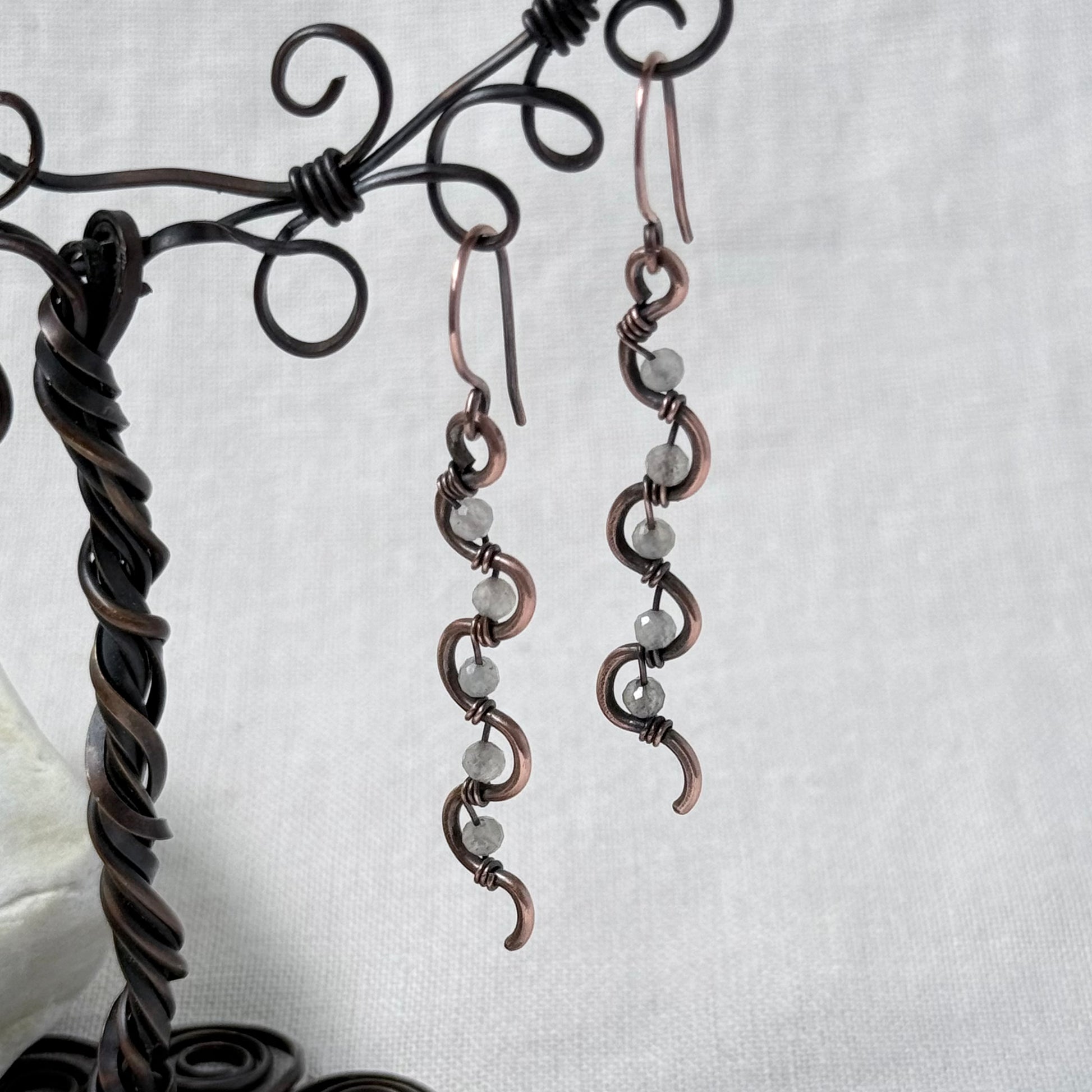A pair of handmade copper wire drop earrings with small Labradorite faceted beads, displayed on a dark metal wire stand against a light grey fabric background.