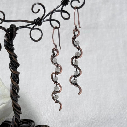 A pair of handmade copper wire drop earrings with small Labradorite faceted beads, displayed on a dark metal wire stand against a light grey fabric background.