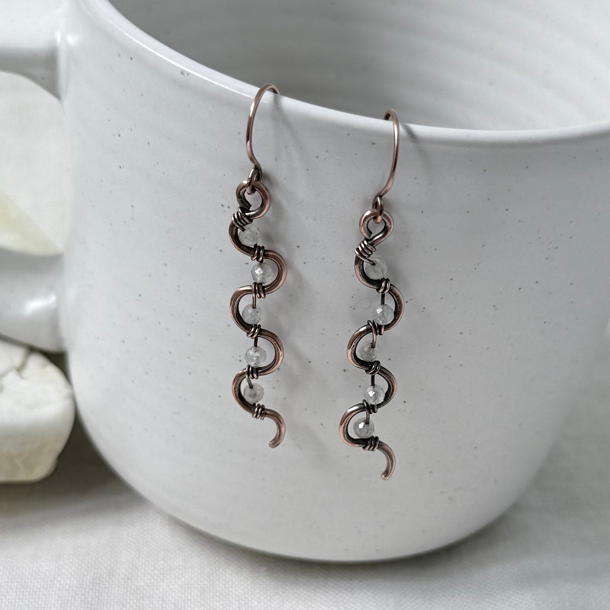 A pair of handmade copper wire dangle earrings with small Labradorite pearl-like beads, hanging from the rim of a white speckled ceramic mug.