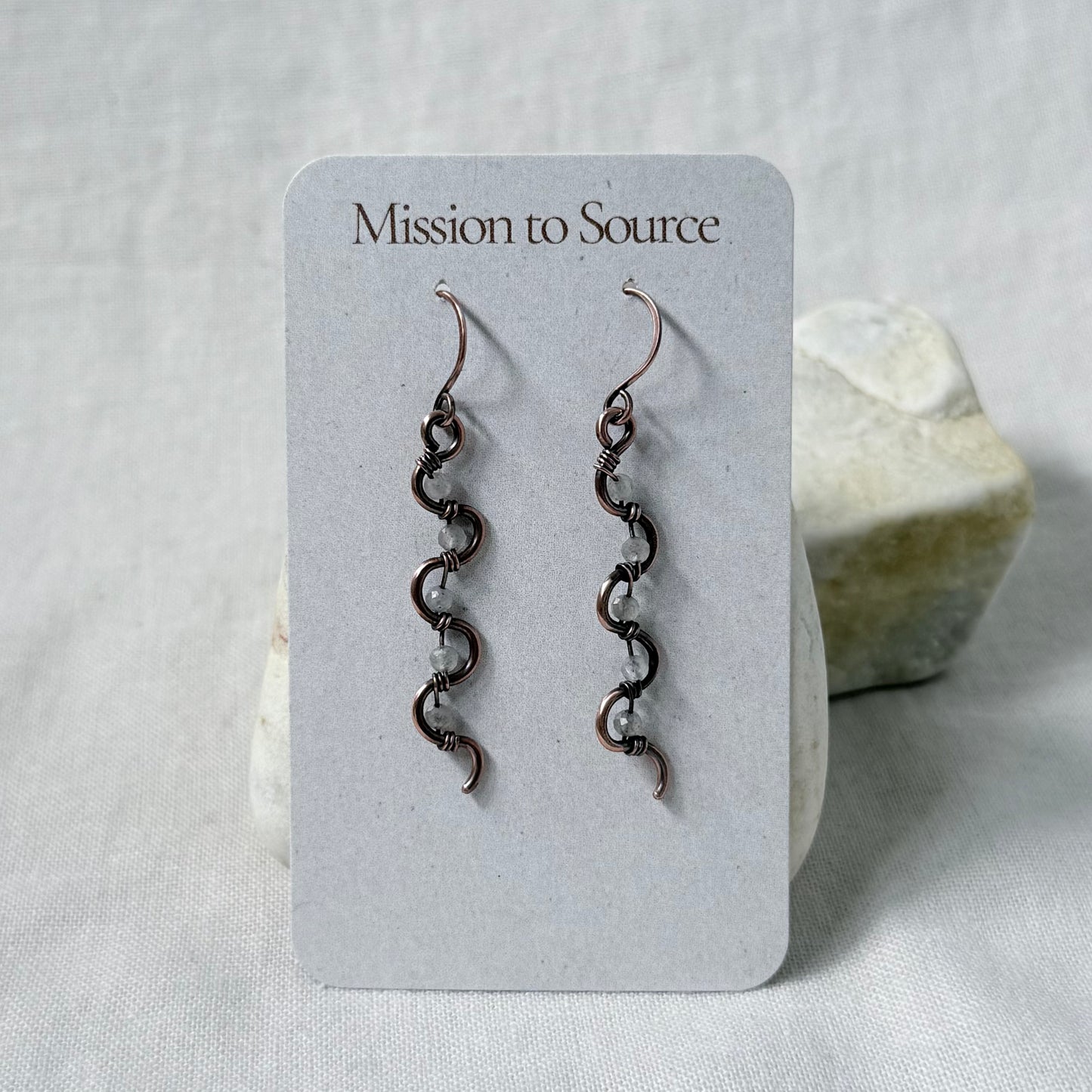 A pair of long, copper dangling earrings with pale Labradorite beads, displayed on a white card labeled 'Mission to Source,' set against a soft white fabric background with a small stone prop.