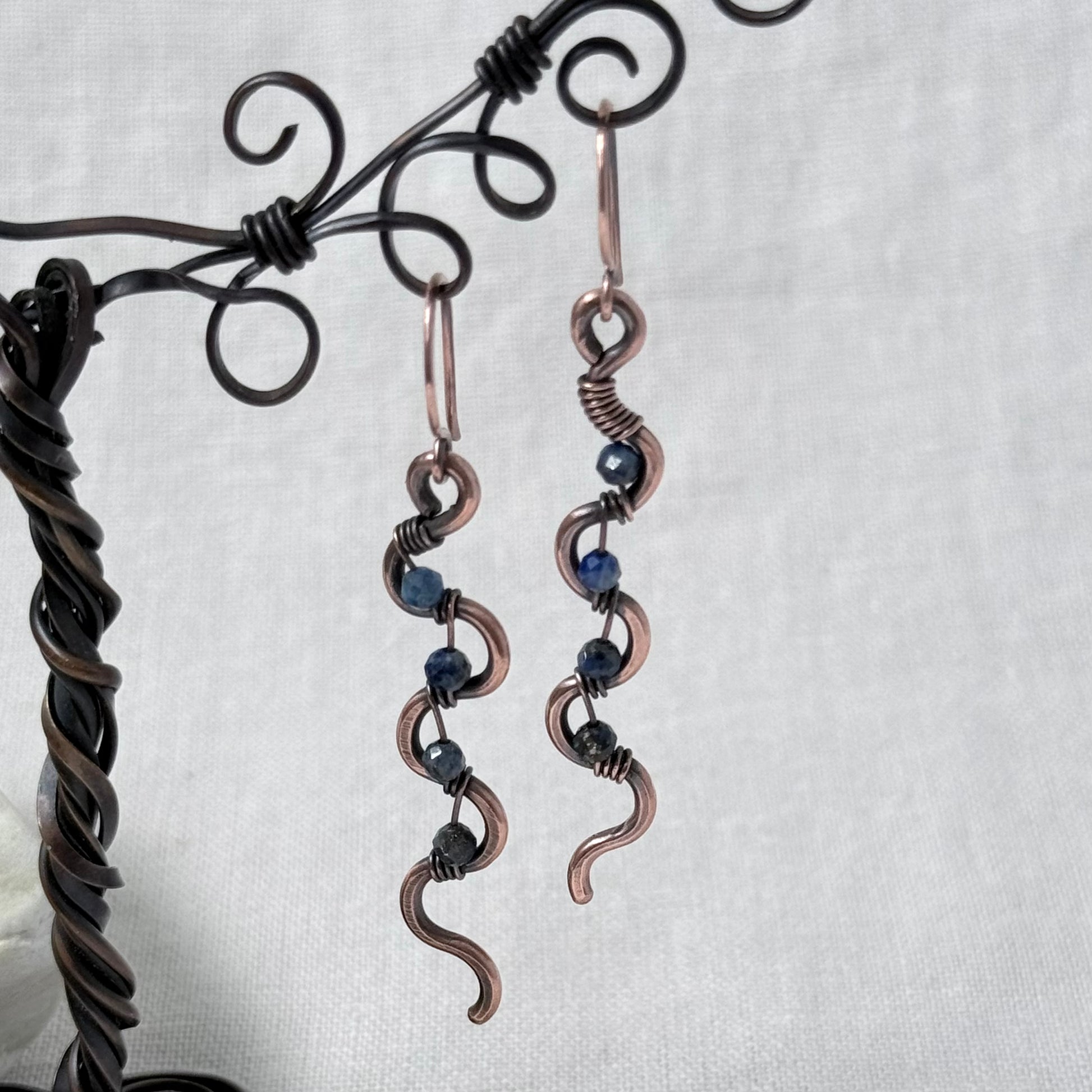 A pair of handmade copper wire dangle earrings with dark blue Lapis Lazuli beads, displayed on a black metal stand against a light grey fabric background.