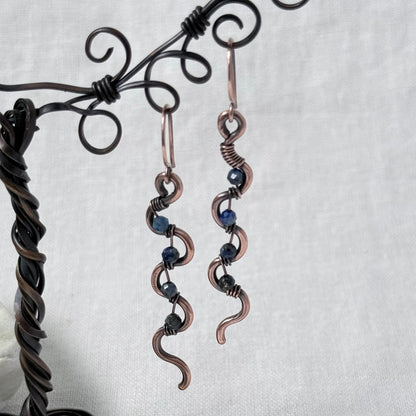 A pair of handmade copper wire dangle earrings with dark blue Lapis Lazuli beads, displayed on a black metal stand against a light grey fabric background.