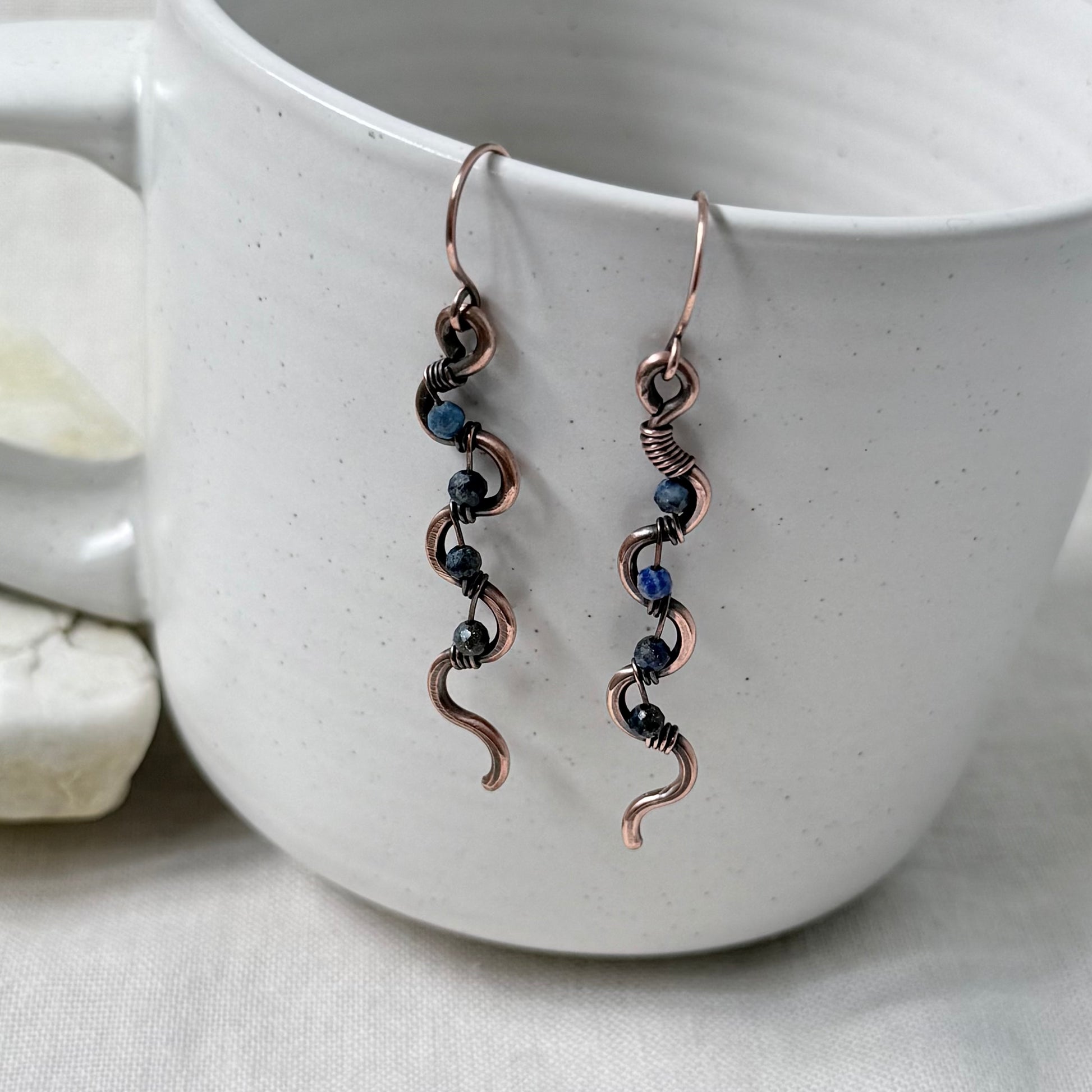 A pair of handmade copper wire dangle earrings with small blue beads, hanging from the rim of a white speckled ceramic mug.