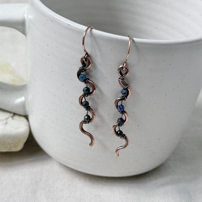 A pair of handmade copper wire dangle earrings with small blue beads, hanging from the rim of a white speckled ceramic mug.