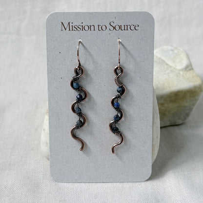 A pair of handmade copper dangle earrings with blue Lapis Lazuli beads, displayed on a white card labeled 'Mission to Source.' 