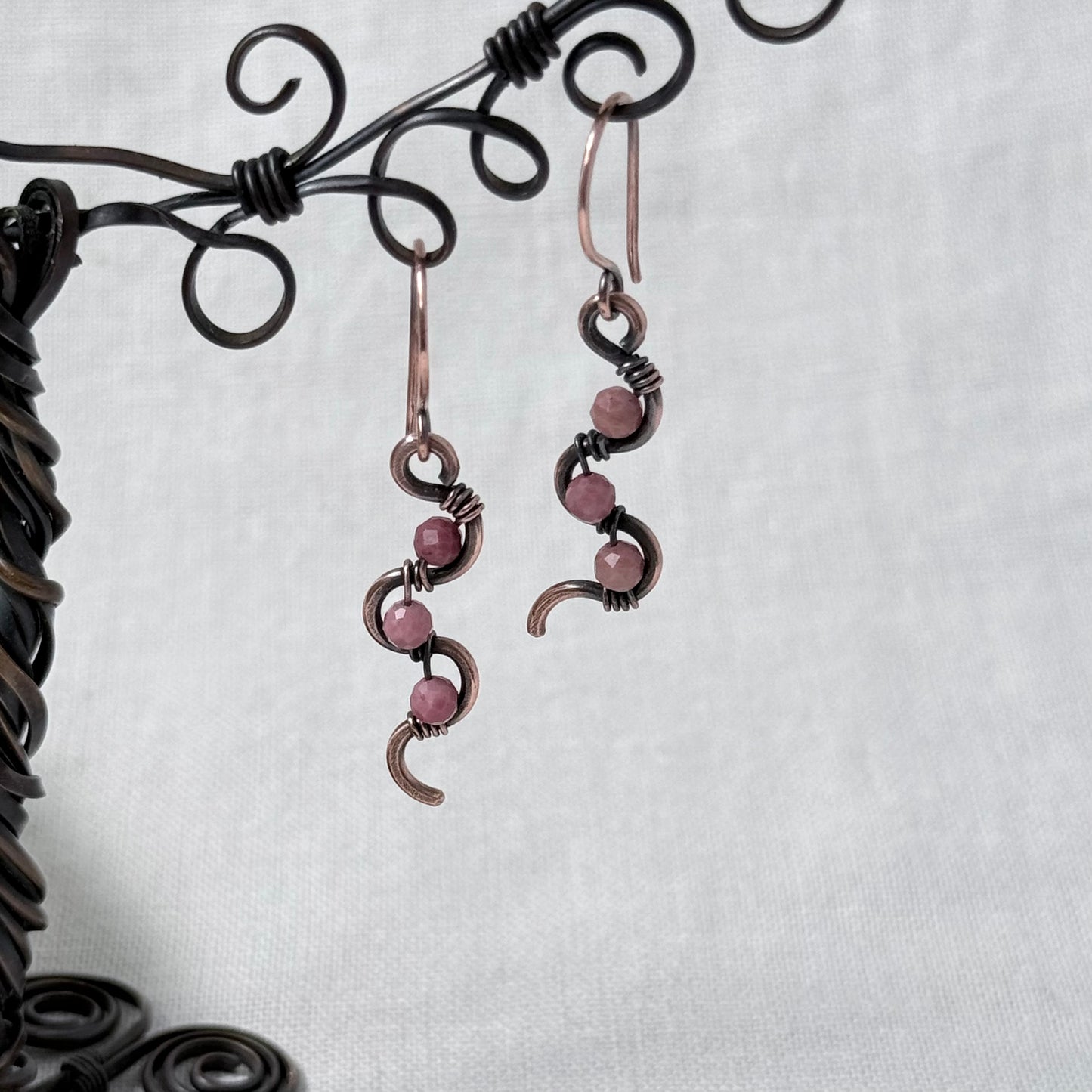 A pair of handmade wire-wrapped drop earrings with pink Rhodonite beads and dark metal accents, hanging from a black decorative display stand against a light grey fabric background.