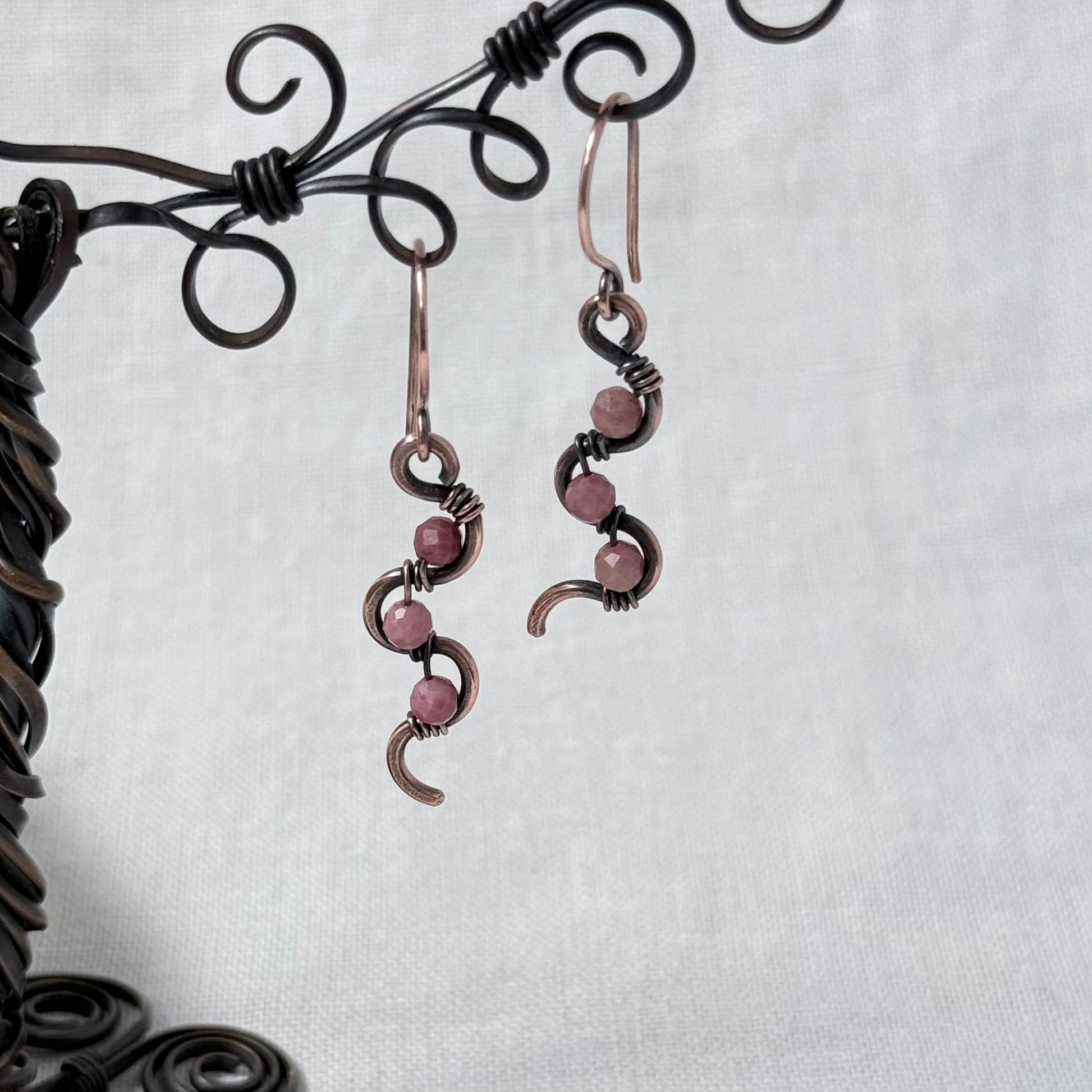 A pair of handmade wire-wrapped drop earrings with pink Rhodonite beads and dark metal accents, hanging from a black decorative display stand against a light grey fabric background.