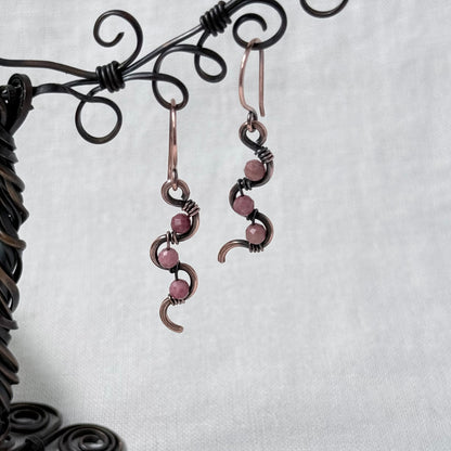 A pair of handmade wire-wrapped drop earrings with pink Rhodonite beads and dark metal accents, hanging from a black decorative display stand against a light grey fabric background.