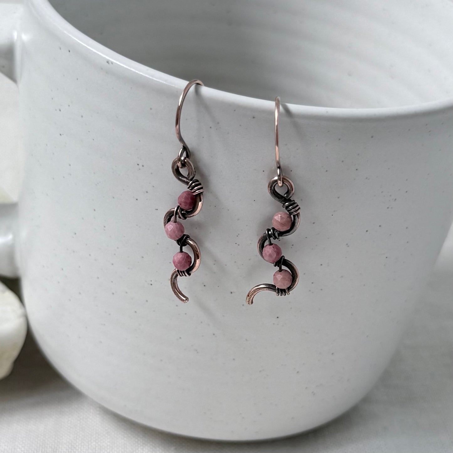 A pair of handmade copper dangle earrings with pink Rhodonite beads, hanging from a white speckled ceramic mug.