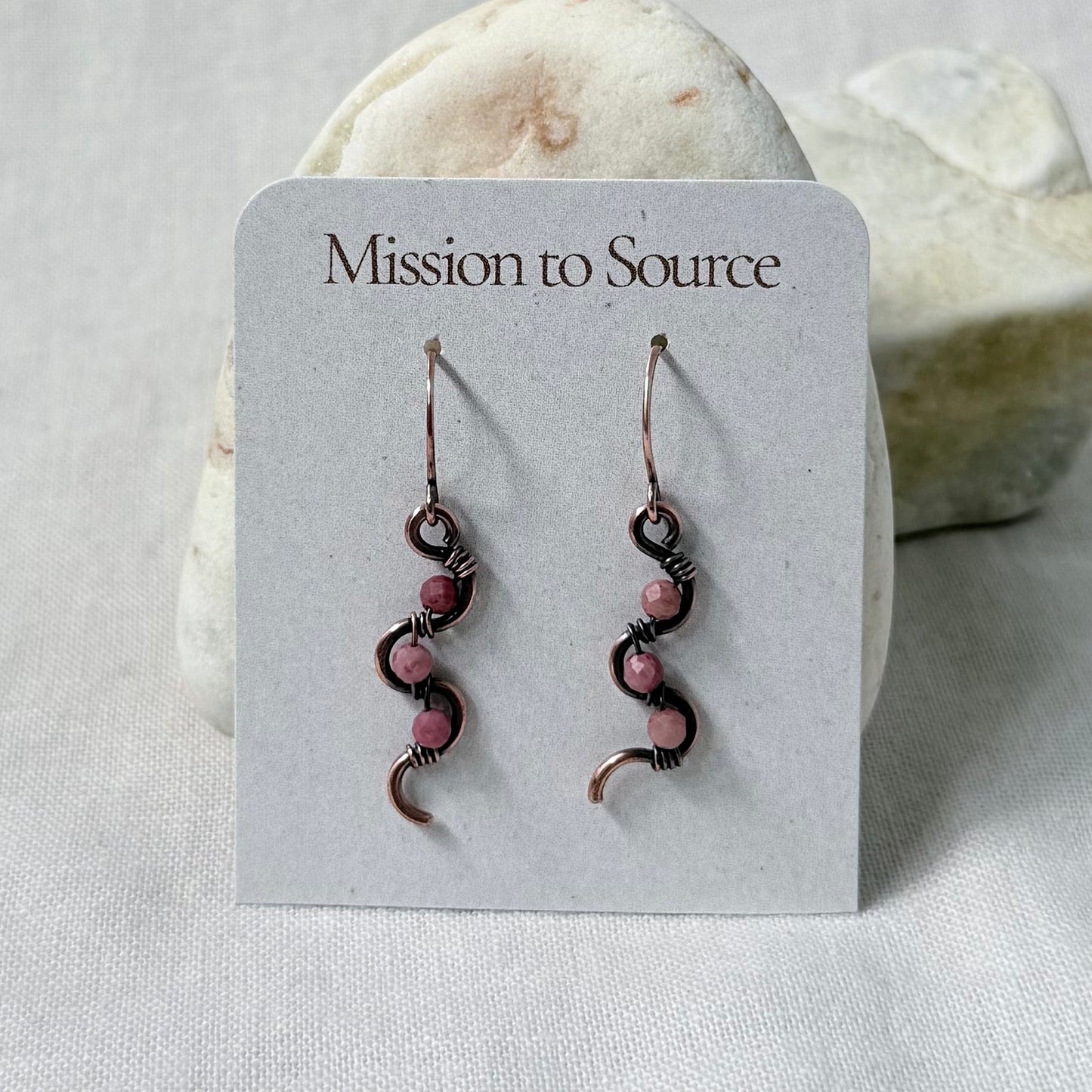 A pair of handmade copper dangle earrings with pink Rhodonite beads, displayed on a white card labeled 'Mission to Source' against a soft-focus stone background.