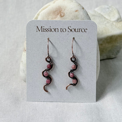 A pair of handmade copper dangle earrings with pink Rhodonite beads, displayed on a white card labeled 'Mission to Source' against a soft-focus stone background.