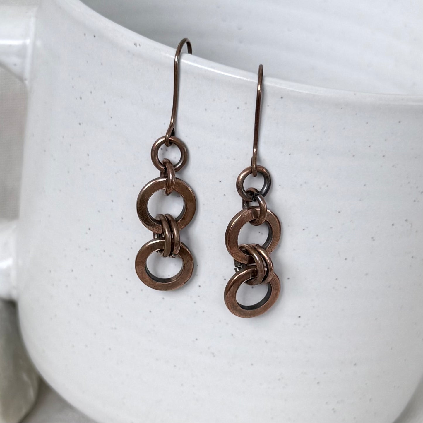 A pair of dangling earrings with interlocking circular copper link, hanging from matching hooks against a white speckled ceramic mug.