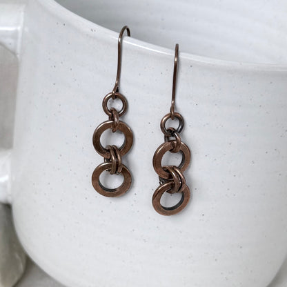 A pair of dangling earrings with interlocking circular copper link, hanging from matching hooks against a white speckled ceramic mug.