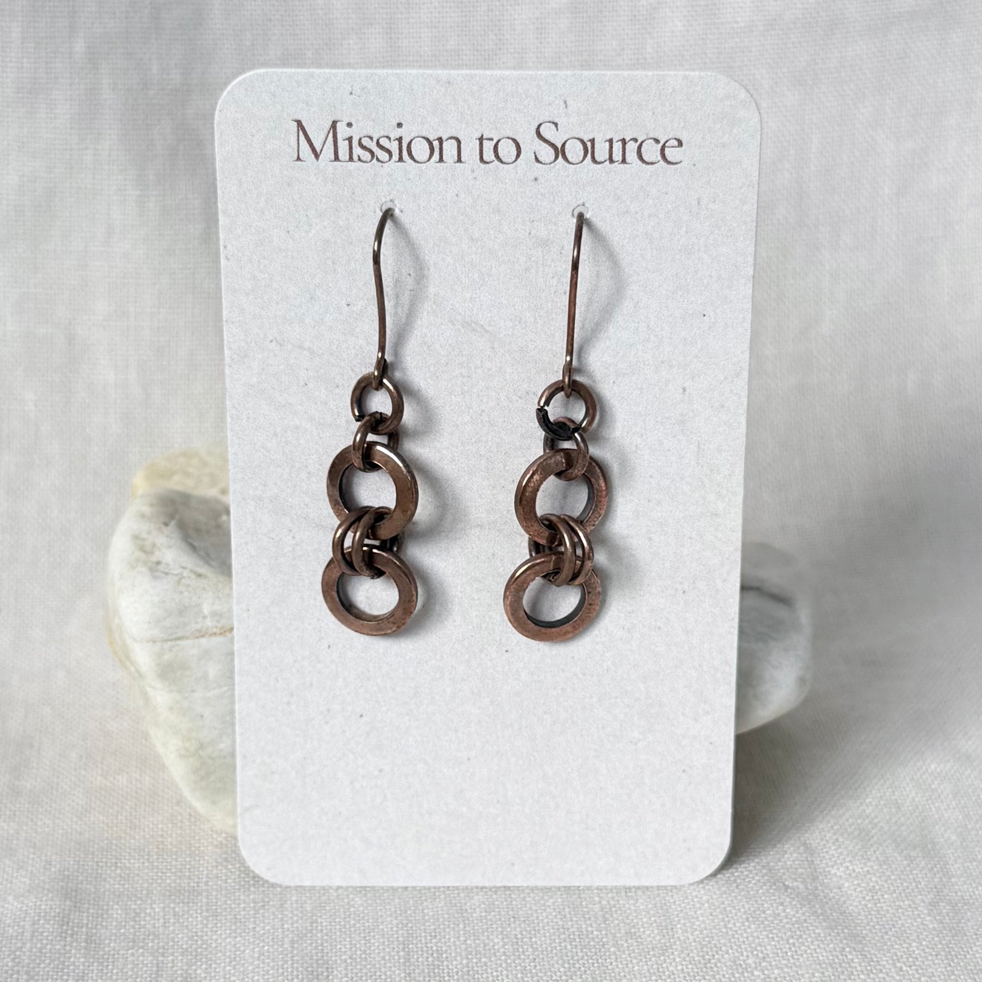 A pair of dangling copper earrings with interlocking circular links, displayed on a white card labeled 'Mission to Source' against a soft neutral fabric background.