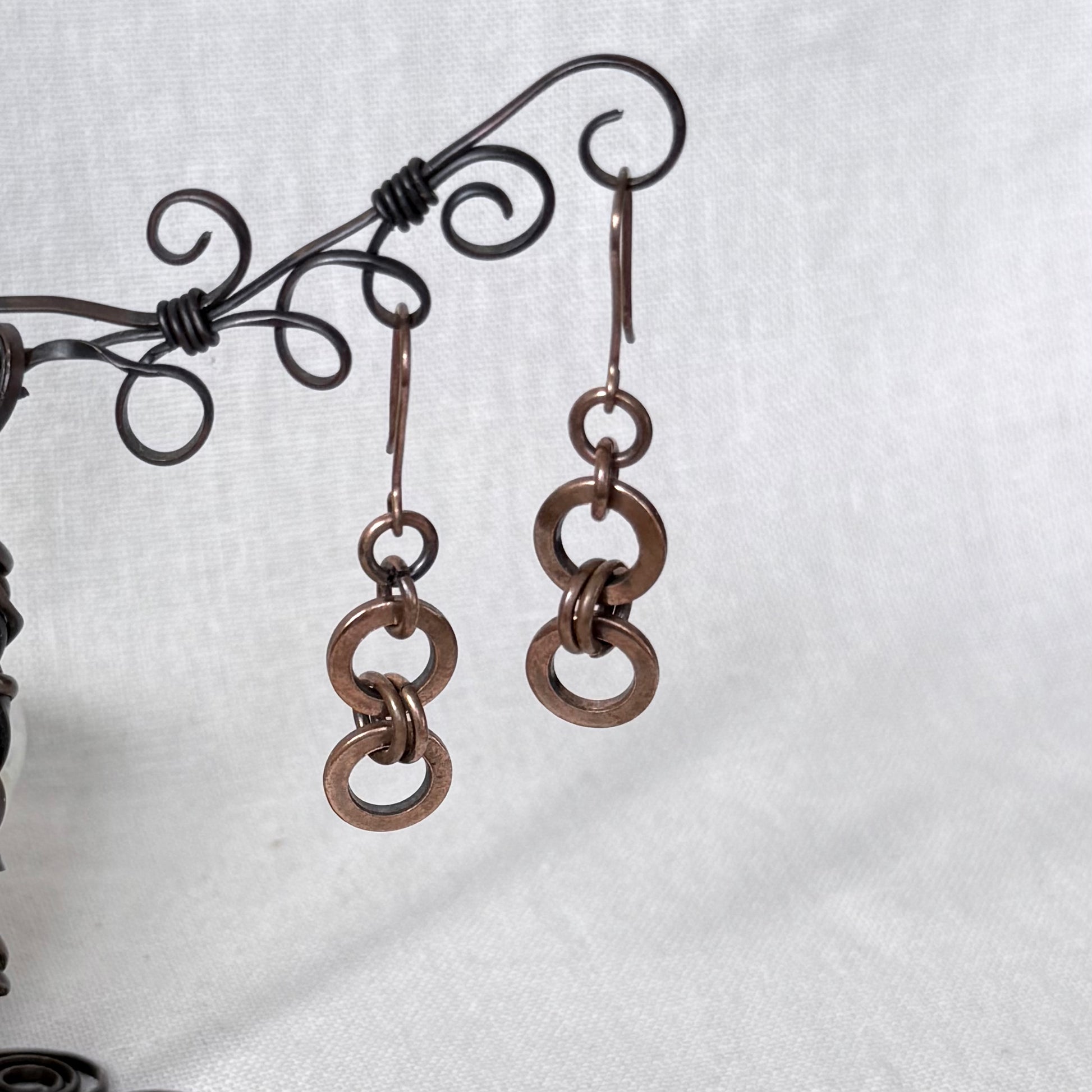 A pair of handmade copper dangle earrings with three interlinked circular links, hanging from a black wire jewellery stand against a white fabric background.