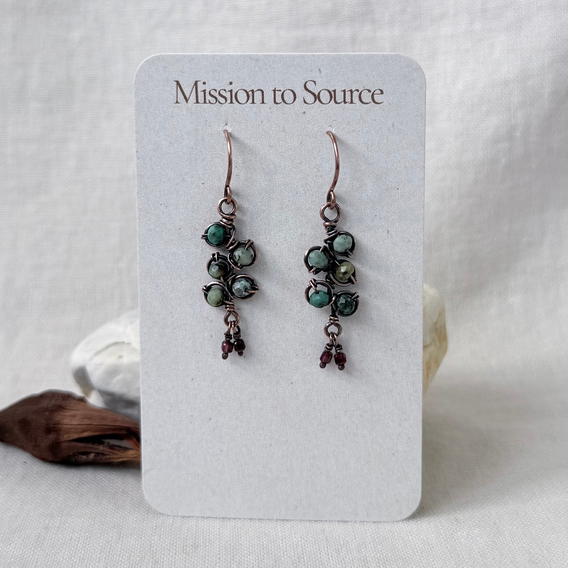 A pair of handmade copper dangle earrings with green marbled beads and dark red bead accents, displayed on a white card labeled 'Mission to Source'.