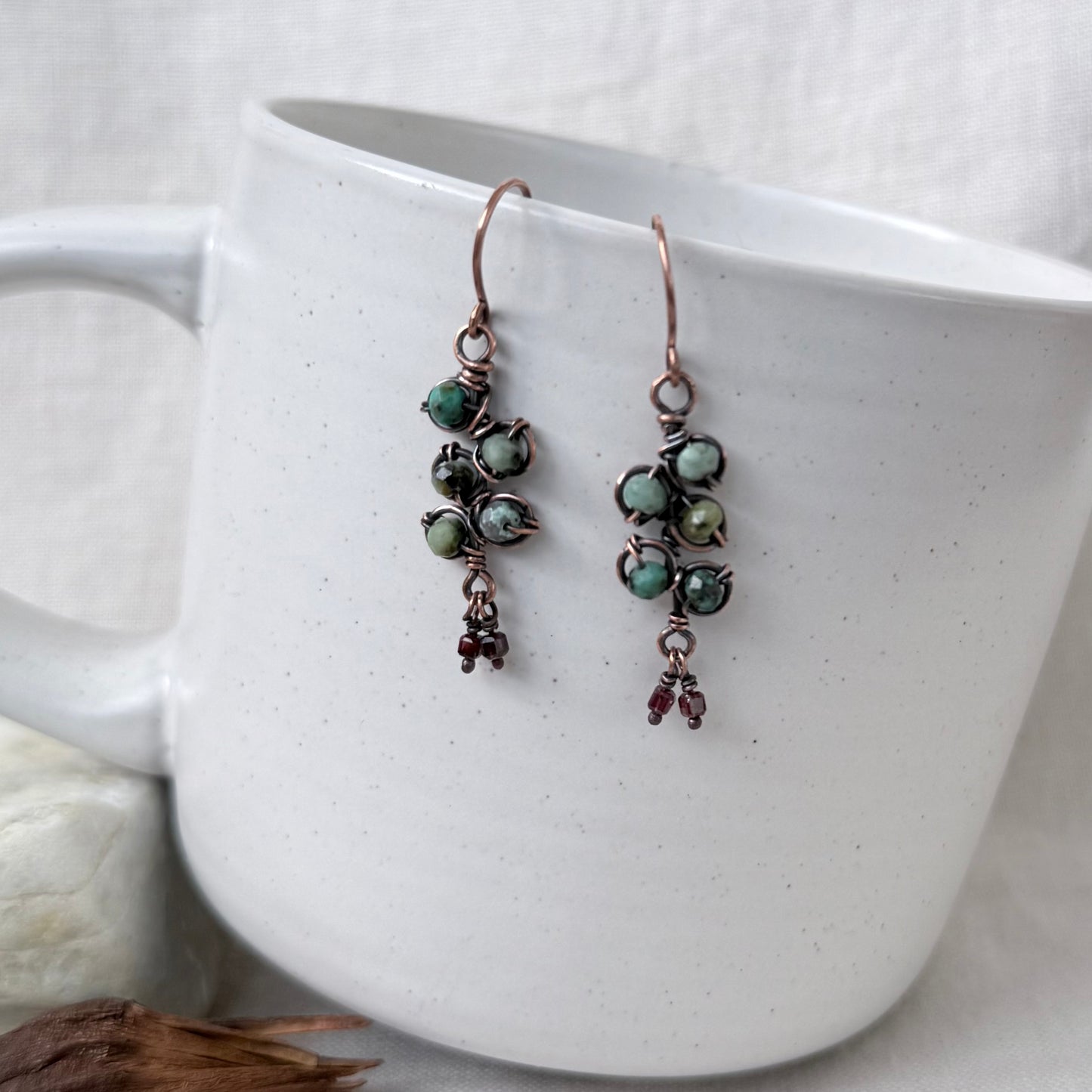 A pair of handmade copper wire drop earrings with green marbled beads and small dark red dangling charms, displayed on the side of a white ceramic mug.