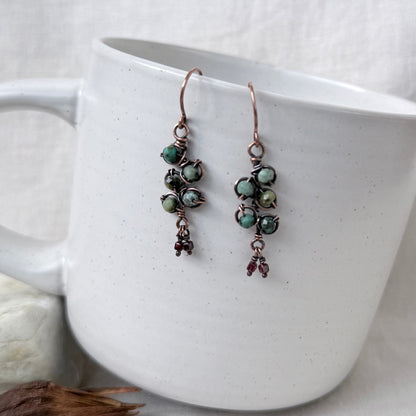 A pair of handmade copper wire drop earrings with green marbled beads and small dark red dangling charms, displayed on the side of a white ceramic mug.