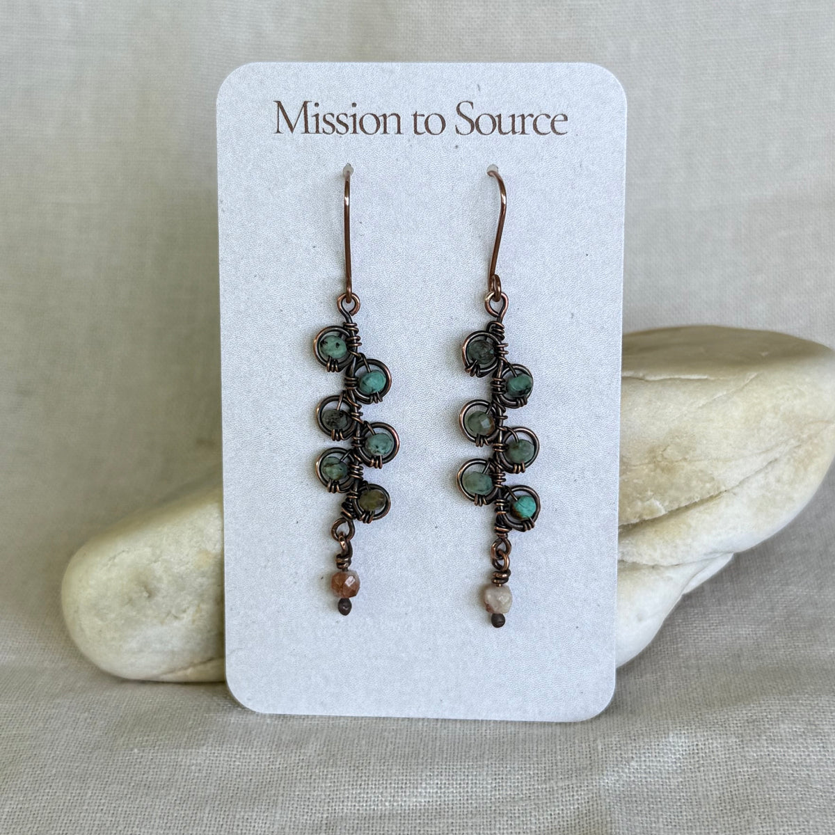 Pair of copper and gemstone earrings on a 'Mission to Source' card with stones in the background
