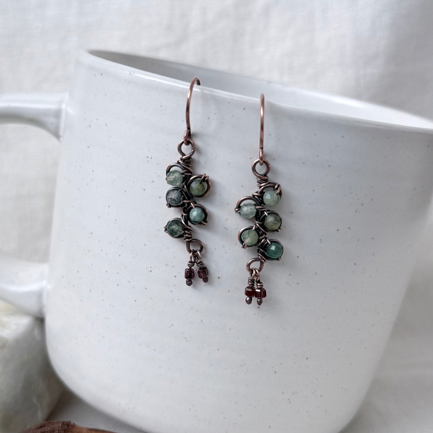 A pair of handmade dangle earrings with green beads and copper wire, hanging from the rim of a white speckled ceramic mug.