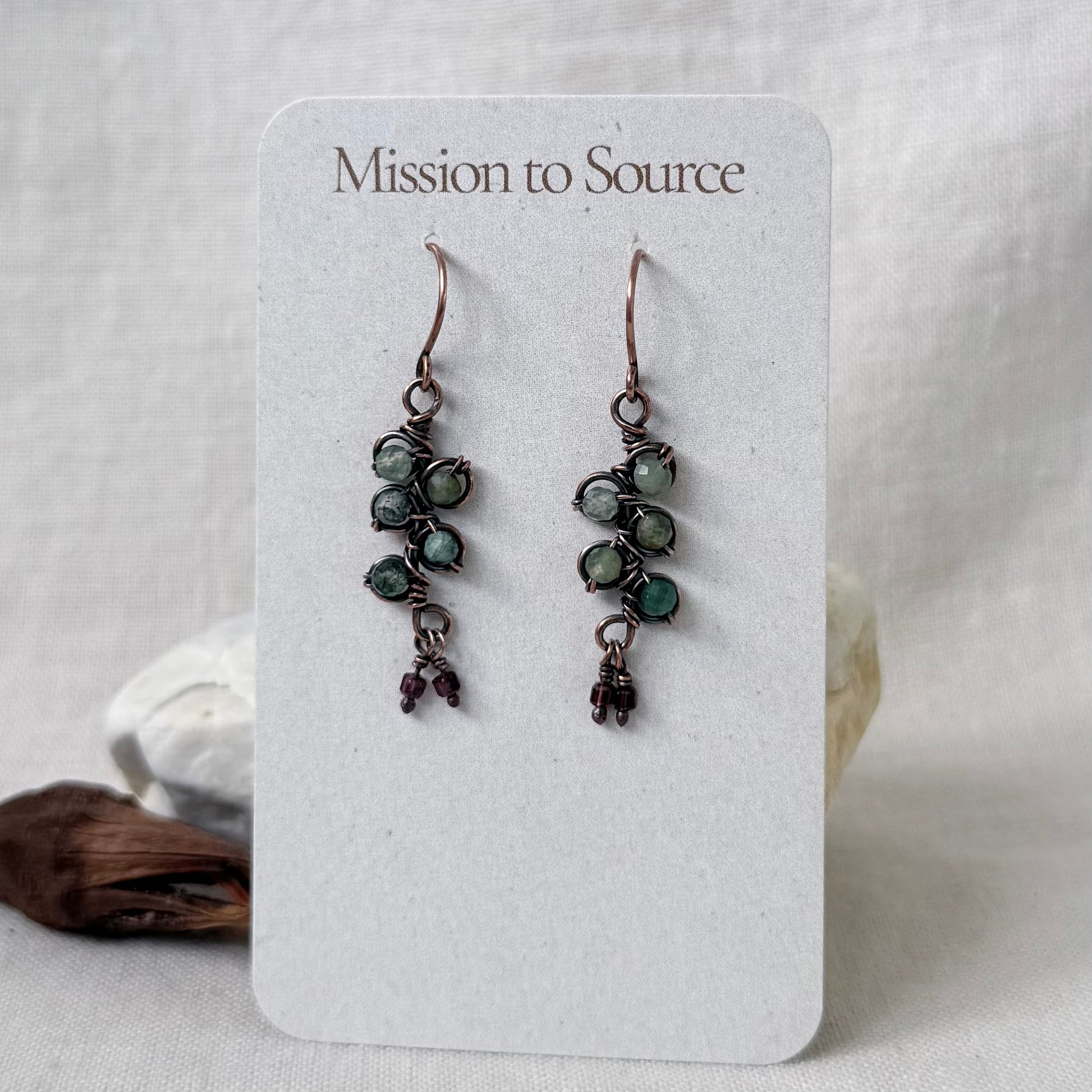 A pair of handmade copper dangle earrings displayed on a white card labeled 'Mission to Source.' The earrings have clusters of small green beads with tiny dangling burgundy accents.