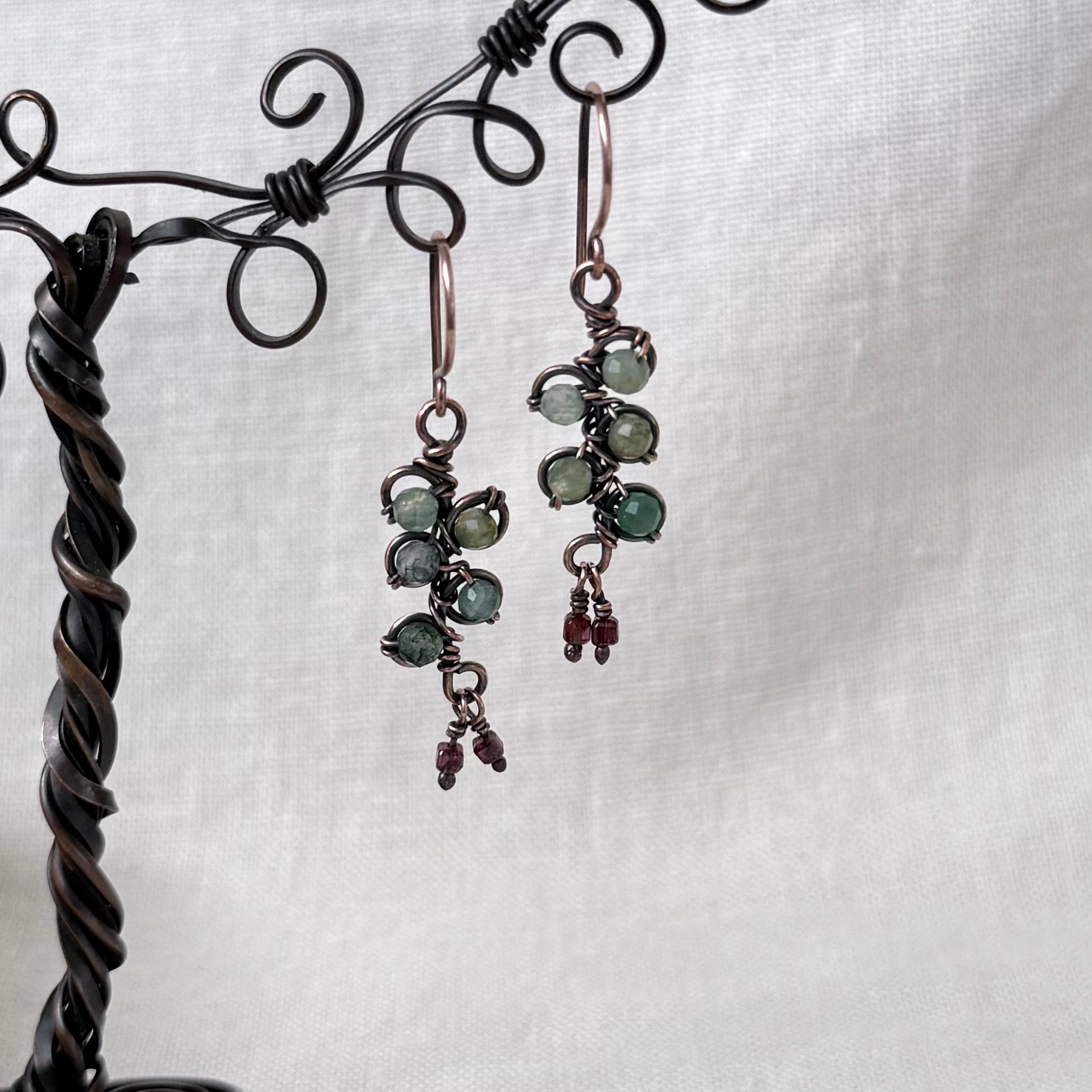 A pair of handmade copper wire-wrapped drop earrings with green gemstone clusters and small red bead accents, hanging from a dark metal display stand against a white textured fabric background.