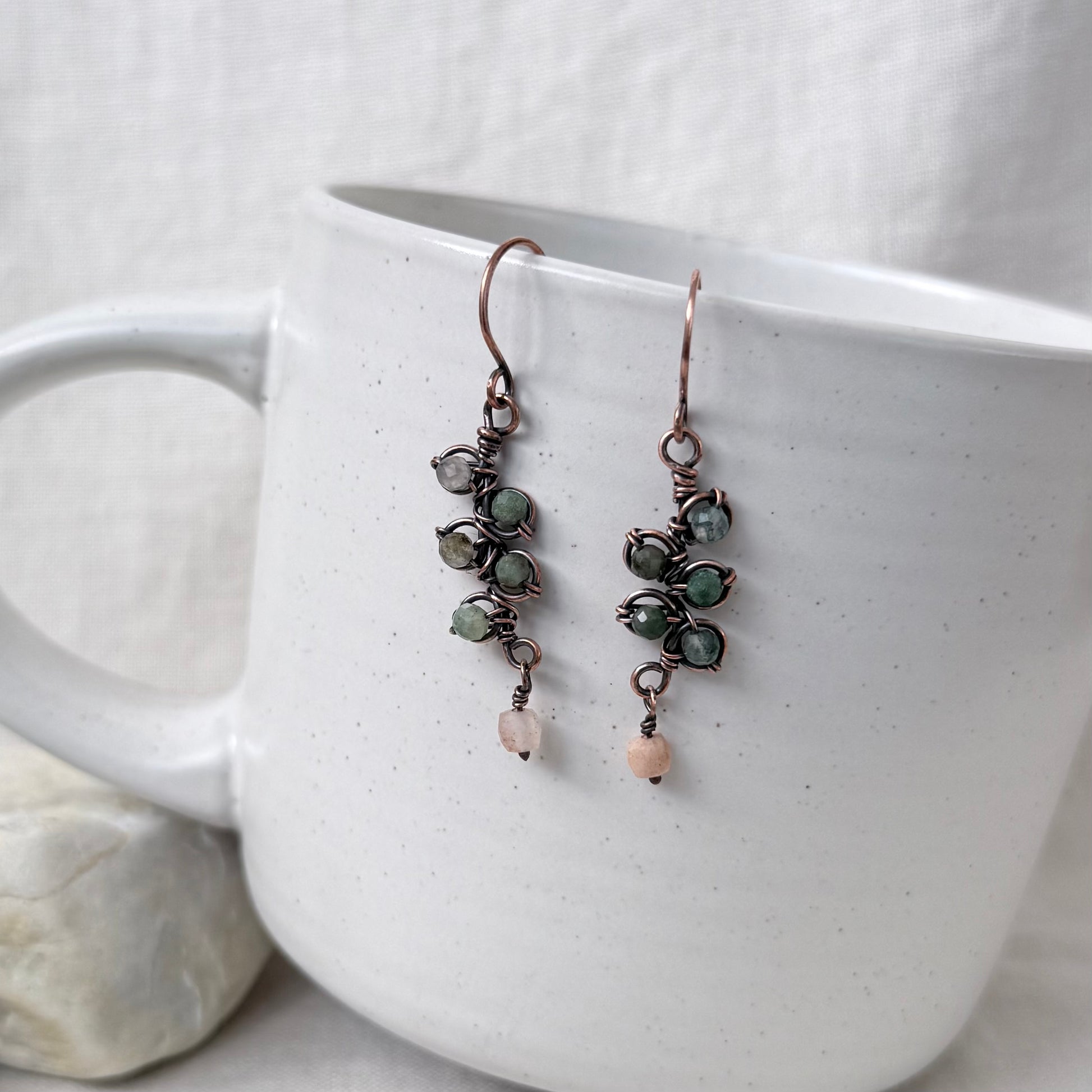 A pair of handmade dangle earrings with copper wire wrapping, green stone clusters, and small peach beads, hanging from the rim of a white ceramic mug.