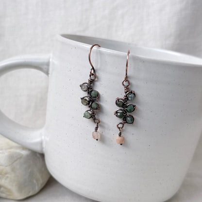 A pair of handmade dangle earrings with copper wire wrapping, green stone clusters, and small peach beads, hanging from the rim of a white ceramic mug.