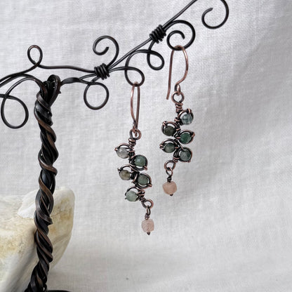 A pair of handmade copper dangle earrings with green and pink gemstones, hanging from a black wire display stand against a white fabric background.